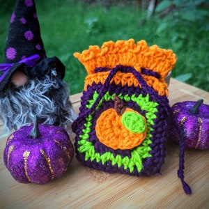 May include: A crocheted pumpkin-shaped bag with green and orange accents. The bag is tied with a purple string and is sitting on a wooden surface next to a purple glitter pumpkin and a gnome wearing a black and purple hat.