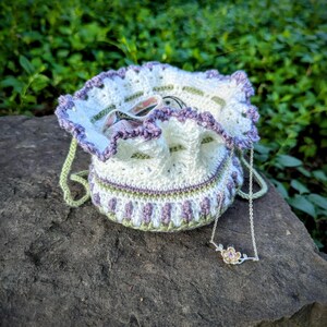 May include: A handmade crochet drawstring bag in white, lavender, and green, with a matching necklace featuring a flower pendant. The bag has a ruffled edge and a drawstring closure. The necklace is silver-toned.