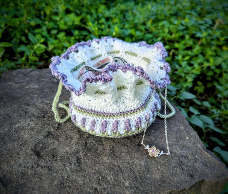 May include: A handmade crochet drawstring bag in white, lavender, and green, with a matching necklace featuring a flower pendant. The bag has a ruffled edge and a drawstring closure. The necklace is silver-toned.