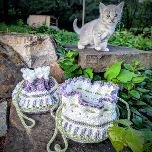 May include: Two handmade crochet drawstring bags in white, lavender, and green, with a matching kitten perched on a stone wall. The bags have a scalloped edge and a detailed pattern. The kitten is gray with blue eyes.