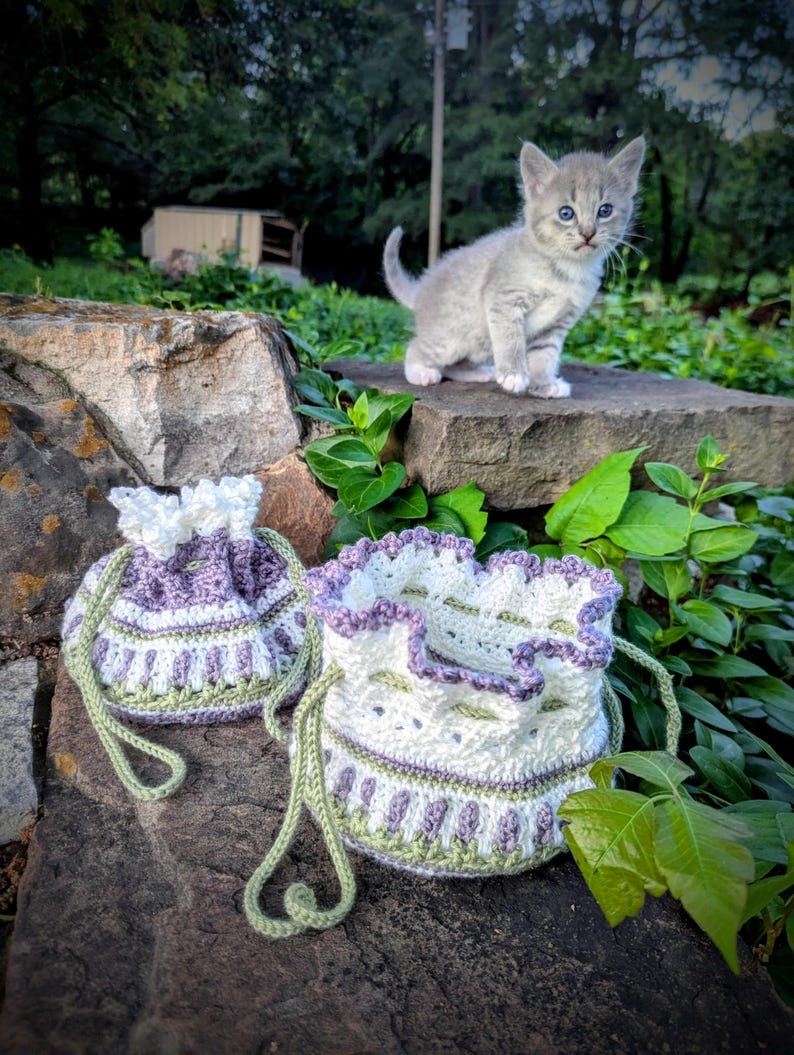 May include: Two handmade crochet drawstring bags in white, lavender, and green, with a matching kitten perched on a stone wall. The bags have a scalloped edge and a detailed pattern. The kitten is gray with blue eyes.