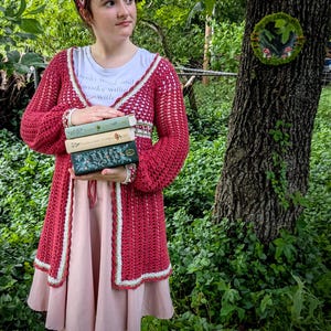 May include: A red crochet cardigan with white trim is worn over a pink dress. The person is holding a stack of books. A white t-shirt with text is visible. The background is a lush green forest.