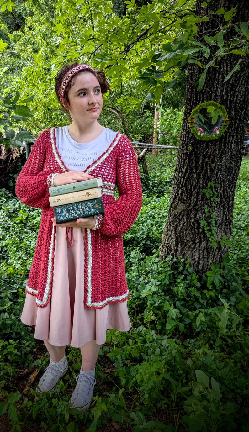 May include: A red crochet cardigan with white trim is worn over a pink dress. The person is holding a stack of books. A white t-shirt with text is visible. The background is a lush green forest.