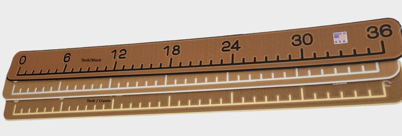 May include: Three brown rulers with black markings. The top ruler has the numbers 0 to 36 printed on it. The middle ruler has white markings and the bottom ruler has gold markings. The top ruler has the text "Teak/Black" printed on it. The middle ruler has the text "Teak/Cream" printed on it. The top ruler has the text "Made in U.S.A." printed on it.