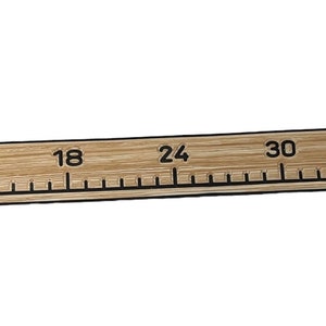Pu&ograve; includere: Un lungo righello piatto in legno chiaro con segni e numeri neri stampati che vanno da 0 a 48 pollici. Il righello ha una consistenza liscia simile alla venatura del legno e include una piccola icona rettangolare della bandiera USA vicino al segno 48.