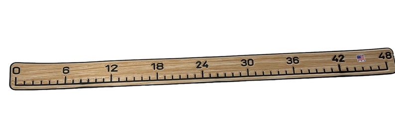 Pu&ograve; includere: Un lungo righello piatto in legno chiaro con segni e numeri neri stampati che vanno da 0 a 48 pollici. Il righello ha una consistenza liscia simile alla venatura del legno e include una piccola icona rettangolare della bandiera USA vicino al segno 48.