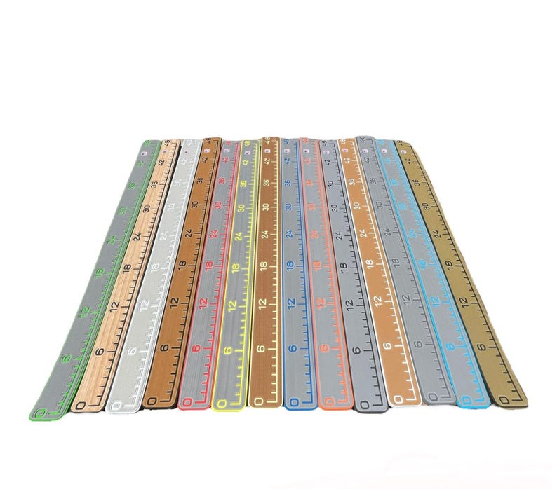 Pu&ograve; includere: Una collezione di dodici righelli rettangolari disposti fianco a fianco a ventaglio. Ogni righello presenta un colore distinto, tra cui verde, beige, bianco, marrone, grigio e blu, con marcature numeriche che vanno da 0 a 106 cm.