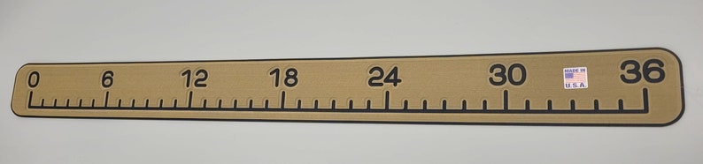 May include: A tan-colored ruler with black markings and numbers. The ruler measures 36 inches and has markings for every inch. The ruler is made in the USA.