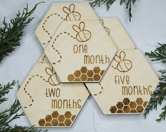 May include: Set of five wooden hexagonal baby milestone cards with a bee and honeycomb design. The cards feature the text "one month", "two months", "five months".
