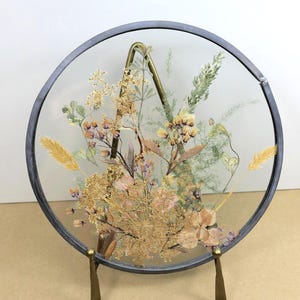 May include: A circular glass frame with a dark metal border containing an arrangement of pressed dried flowers in shades of beige, yellow, and muted green. The frame is supported by a brass-toned metal stand with two front legs and a back support.