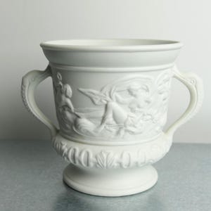 May include: A matte white ceramic urn featuring two side handles and a relief sculpture depicting figures in motion. The base is decorated with a repeating leaf pattern, and the vessel has a wide, flared rim and a circular pedestal foot.