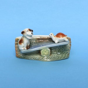 May include: A ceramic planter shaped like a wall with a dog and a turtle on a seesaw. The planter is green, brown, and white.