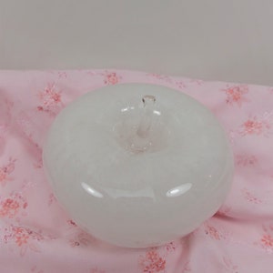 May include: A white glass apple shaped paperweight. The apple is sitting on a pink fabric with a floral pattern.