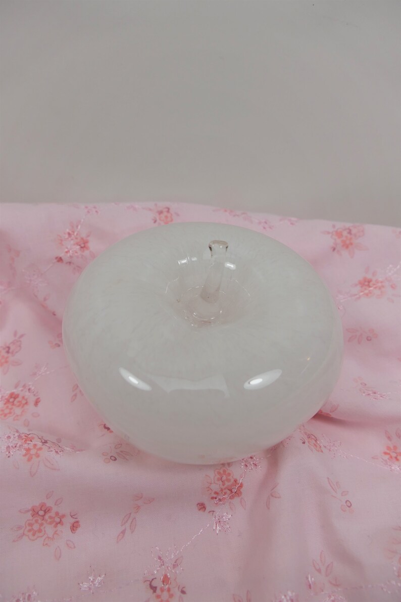 May include: A white glass apple shaped paperweight. The apple is sitting on a pink fabric with a floral pattern.
