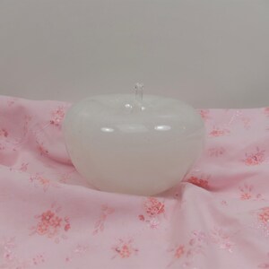 May include: A white glass apple shaped bowl with a clear glass stem. The bowl is sitting on a pink fabric with a floral pattern.