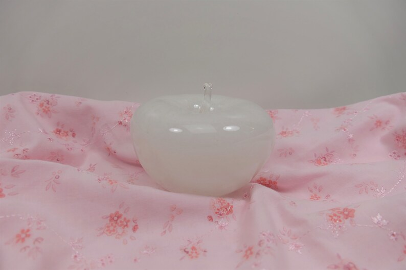 May include: A white glass apple shaped bowl with a clear glass stem. The bowl is sitting on a pink fabric with a floral pattern.