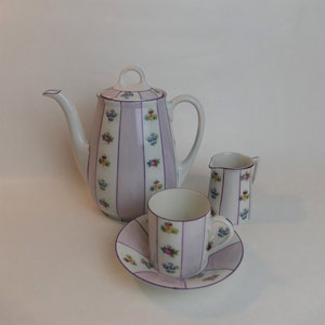 May include: A white tea set with a purple and floral design. The set includes a teapot, a creamer, a cup, and a saucer.