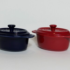 Puede incluir: Dos pequeñas cazuelas de cerámica ovaladas con tapas. Una es azul y la otra es roja.