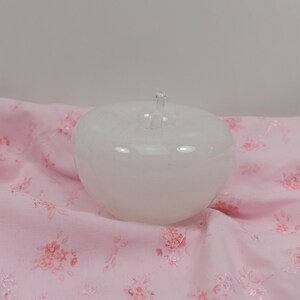 May include: A white glass apple shaped object with a clear glass stem. The object is sitting on a pink fabric with a floral pattern.
