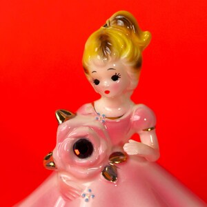 May include: A porcelain figurine of a girl in a pink dress holding a pink rose. The girl has blonde hair and blue eyes. The figurine is in a vintage style.