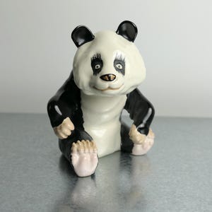 May include: A ceramic panda figurine featuring a glossy white and black finish. The panda is depicted in a seated position with its front paws resting near its feet. It has painted black eye patches, a small black nose, and light-colored paw pads.
