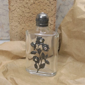 May include: A clear glass bottle with a silver stopper and a silver rose design with rhinestones. The bottle is sitting on a brown paper surface.