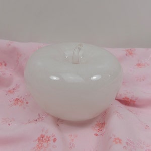 May include: A white glass apple figurine with a clear stem. The apple is sitting on a pink fabric with a floral pattern.