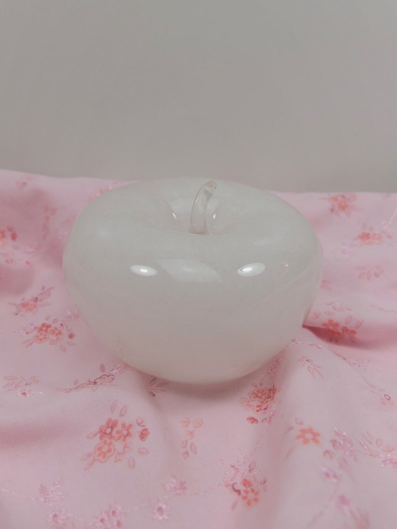 May include: A white glass apple figurine with a clear stem. The apple is sitting on a pink fabric with a floral pattern.