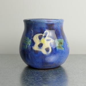 May include: A small, rounded ceramic cup with a glossy finish in shades of deep blue. The exterior features a hand-painted, stylized white flower design with a yellow center, accented by smaller green floral motifs encircling the body.