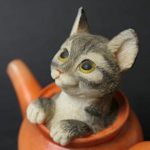 May include: A grey and white tabby kitten figurine peeking out of an orange teapot.