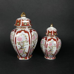 Puede incluir: Dos jarrones de jengibre de porcelana con diseños florales y de pájaros ornamentados. El jarrón más grande es rojo con detalles dorados, mientras que el jarrón más pequeño es de un tono rojo más claro con detalles dorados. Ambos jarrones tienen un fondo blanco.