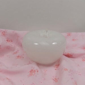 May include: A white ceramic apple shaped candle on a pink floral fabric background.