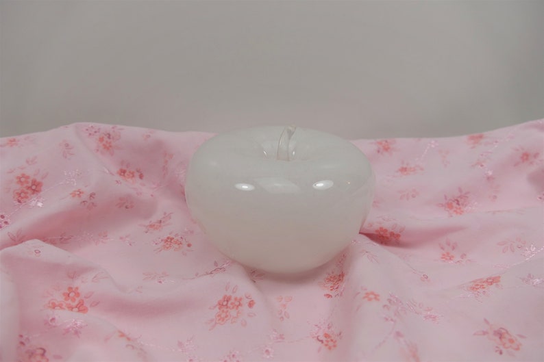 May include: A white ceramic apple shaped candle on a pink floral fabric background.