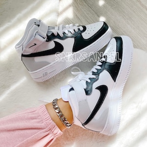 May include: White and black high-top trainers with a black Nike swoosh logo. The trainers have a white midsole and a black outsole. The trainers are on a white fluffy surface.