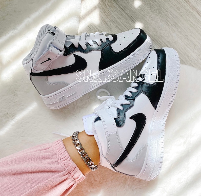 May include: White and black high-top trainers with a black Nike swoosh logo. The trainers have a white midsole and a black outsole. The trainers are on a white fluffy surface.