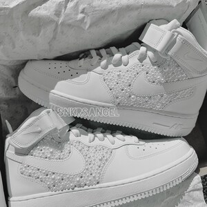 K&ouml;nnte beinhalten: Ein Paar wei&szlig;e High-Top-Sneaker mit einer perlenartigen Verzierung auf dem Swoosh-Logo und den Seitenpaneelen. Die Sneaker haben eine wei&szlig;e Mittelsohle und Au&szlig;ensohle. Der Text "SNKRSANGEL" ist an der Seite des Schuhs sichtbar.