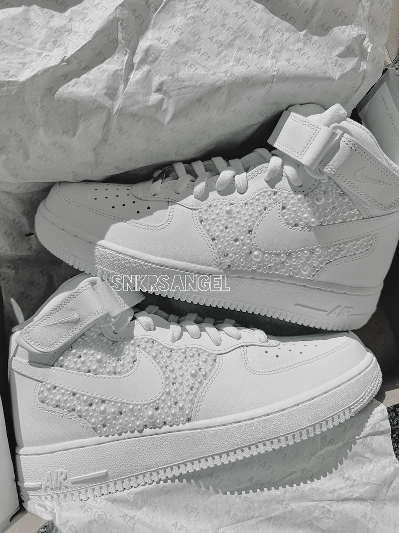 K&ouml;nnte beinhalten: Ein Paar wei&szlig;e High-Top-Sneaker mit einer perlenartigen Verzierung auf dem Swoosh-Logo und den Seitenpaneelen. Die Sneaker haben eine wei&szlig;e Mittelsohle und Au&szlig;ensohle. Der Text "SNKRSANGEL" ist an der Seite des Schuhs sichtbar.