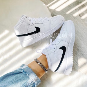 May include: White leather mid-top trainers with a black swoosh and black trim. The trainers are worn by a person wearing blue jeans and a silver chain bracelet.