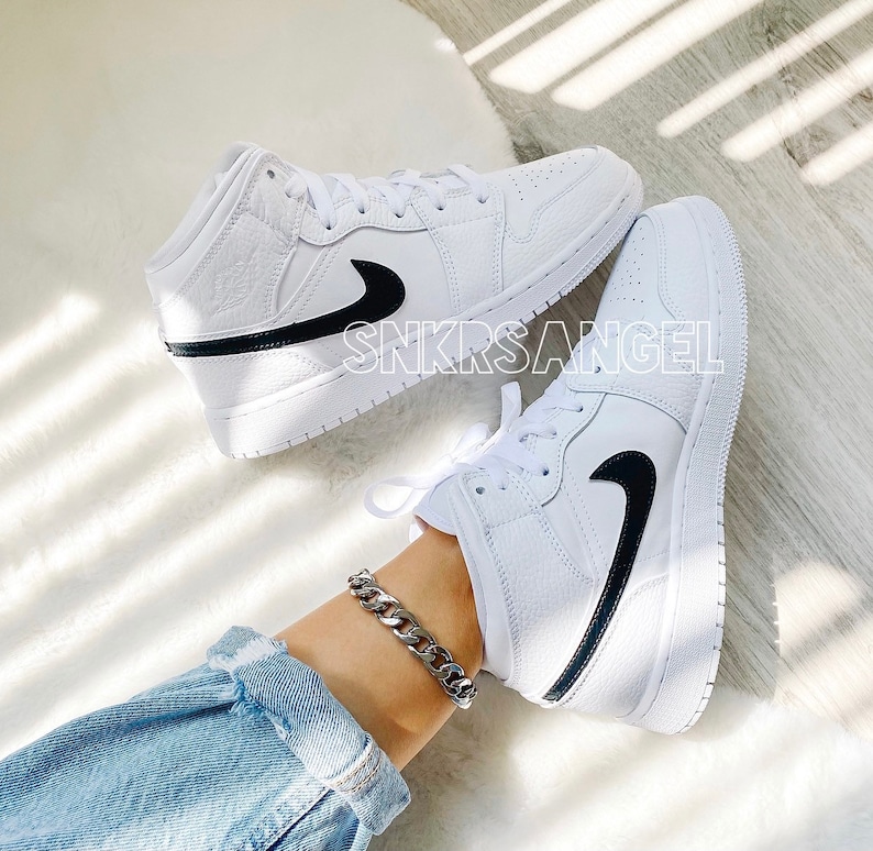 May include: White leather mid-top trainers with a black swoosh and black trim. The trainers are worn by a person wearing blue jeans and a silver chain bracelet.