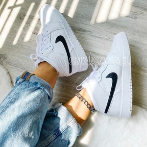 May include: White leather mid-top trainers with a black swoosh logo. The trainers are worn with blue jeans and a silver chain ankle bracelet.