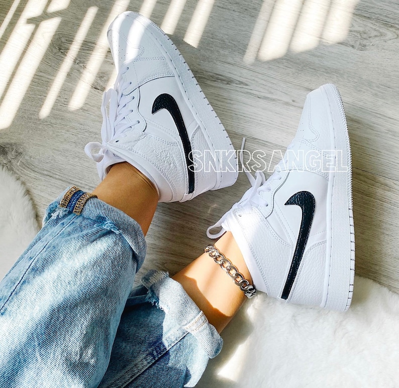 May include: White leather mid-top trainers with a black swoosh logo. The trainers are worn with blue jeans and a silver chain ankle bracelet.