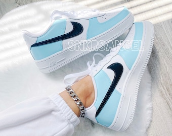 Può includere: Un paio di scarpe Nike Air Force 1 bianche e blu chiaro con swoosh neri. Le scarpe sono indossate da una persona che indossa pantaloni bianchi e un braccialetto a catena argentato.
