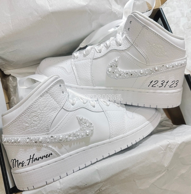 K&ouml;nnte beinhalten: Ein Paar wei&szlig;e High-Top-Sneaker mit Perlenbesatz am Swoosh und den Schn&uuml;rsenkeln. Die Sneaker sind mit dem Text "Mrs. Harren" auf einem Schuh und "12.31.23" auf dem anderen personalisiert.