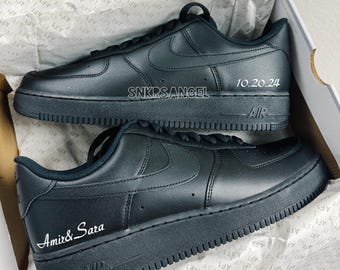 May include: A pair of black leather sneakers with white stitching and a white Nike swoosh logo. The sneakers have the text "SNKRSANGEL" and "10.20.24" on one shoe and "Amir& Sara" on the other shoe.