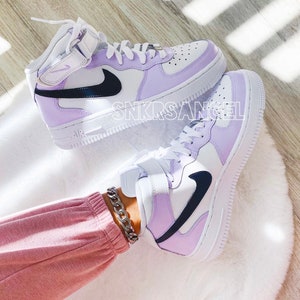 Op de afbeelding: Een paar witte en lavendelkleurige high-top sneakers met zwarte Nike swoosh logo's. De sneakers worden gedragen door iemand die een lavendelkleurige broek en een zilveren kettingarmband draagt.