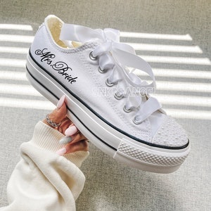 May include: White canvas sneakers with a white satin ribbon tied around the laces. The sneakers have a black embroidered design that says "Mrs. Bride" on the side. The toe of the shoe is decorated with white pearl-like embellishments.