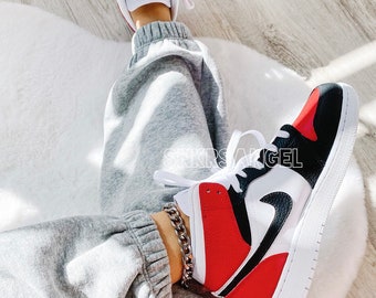May include: A pair of grey sweatpants with a black and red high-top sneaker with a white sole. The sneaker has a white swoosh logo and a silver chain bracelet on the ankle.