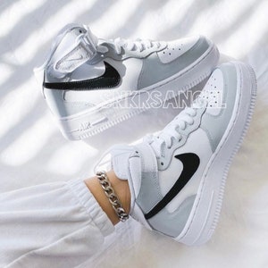 K&ouml;nnte beinhalten: Ein Paar wei&szlig;e High-Top-Sneaker mit schwarzen Nike-Swoosh-Logos. Die Sneaker befinden sich an einem Fu&szlig; einer Person, die ein wei&szlig;es Armband tr&auml;gt.