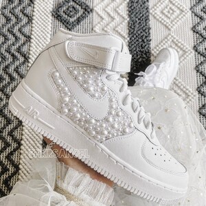 K&ouml;nnte beinhalten: Wei&szlig;e High-Top-Sneaker mit Perlenbesatz an der Seite. Die Sneaker haben ein wei&szlig;es Swoosh-Logo und den Schriftzug "AIR" an der Seite.