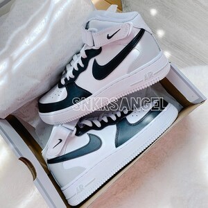 May include: A pair of white high-top trainers with black accents. The trainers have a black swoosh logo on the side and a black toe cap. The trainers are in a box with the word "SNKRSANGEL" printed on the side.